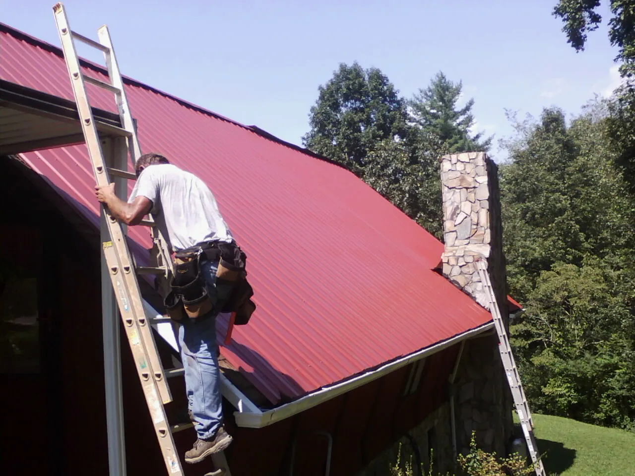 Roofer on a ladder beside a red metal roof and stone chimney — Soffit & Fascia Repair by Roofing Brothers in Manilla